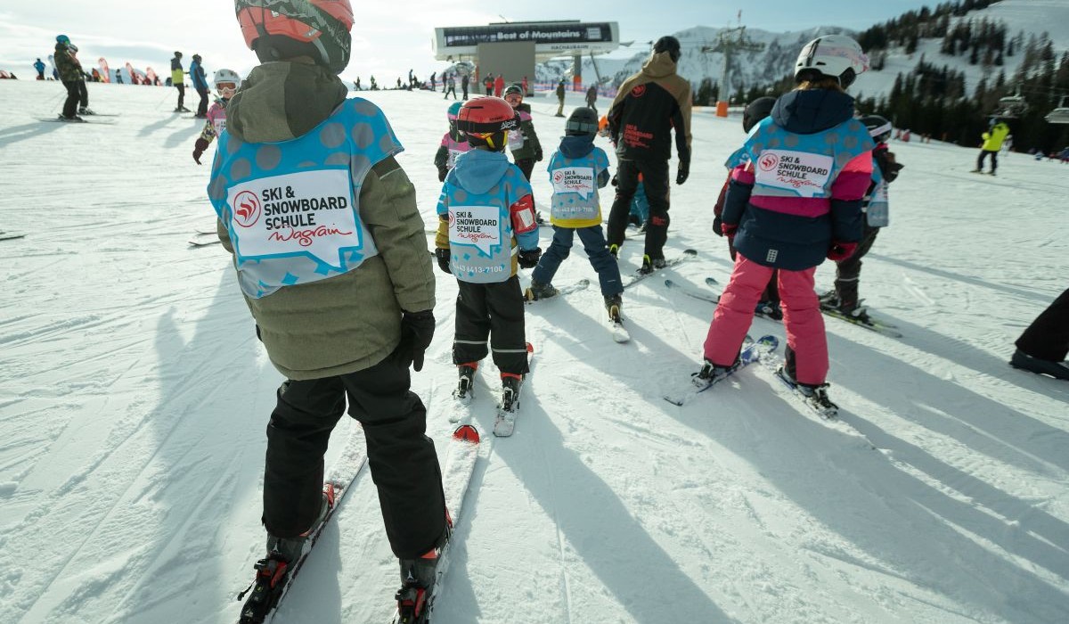 Kinderen in skikleding staan in een skischool op een besneeuwde piste.