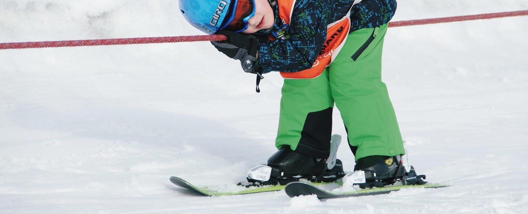 Ein Kind in grüner Skihose zieht mit Skiern an einem roten Seil im Schnee