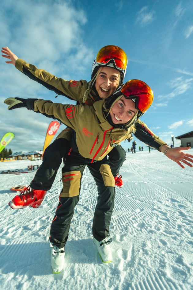 Zwei lachende Menschen beim Wintersport, einer trägt den anderen Huckepack im Schnee
