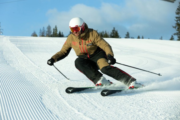 Skifahrer biegt schwungvoll auf präparierter Piste, vor blauem Himmel und Bäumen